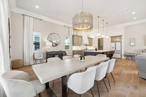 A spacious dining area with a long table and white chairs, a chandelier, and a kitchen in the background.