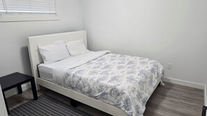 A single bed with a white headboard and floral bedding, a small black nightstand, and a window with blinds.