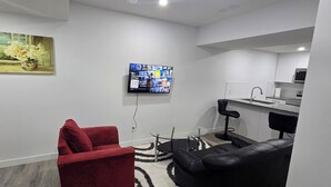 A modern living room with a red sofa, a black leather couch, and a glass coffee table. A flat-screen TV is mounted on the wall, and a painting is visible on the left.