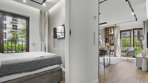 Quarto moderno com cama, escrivaninha e cadeira. Há uma janela com vista para vegetação e uma varanda.