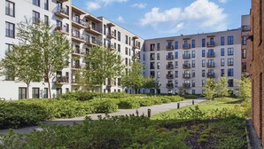 Um complexo de apartamentos moderno com várias varandas, um pátio central com vegetação e um céu claro.