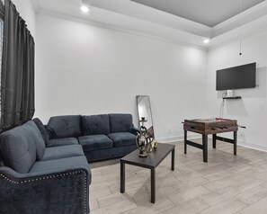 A living room with a dark blue sofa, a coffee table, a foosball table, a mirror, and a flat-screen TV mounted on the wall. - Luxurious Apartment in Houston. (Houston)