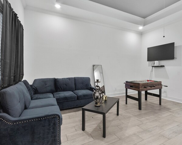 A living room with a dark blue sofa, a coffee table, a foosball table, a mirror, and a flat-screen TV mounted on the wall. - Luxurious Apartment in Houston. (Houston)