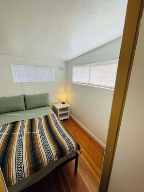 A small, cozy bedroom with a single bed, a nightstand with a lamp, and two windows with blinds.