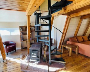 A loft-style room with a wooden interior, a spiral staircase, a wood stove, a bookshelf, and a leather chair.