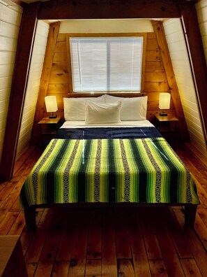 Une chambre confortable située sous les toits, avec un plafond en pente en bois, une grande fenêtre avec des stores, un lit recouvert d’une courtepointe à rayures et deux lampes de chevet.