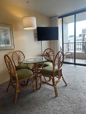 A hotel room with a round glass table, wicker chairs with green cushions, a flat-screen TV mounted on the wall, and a large window with a view of buildings. - Penthouse condo in gorgeous Kihei (Kihei)