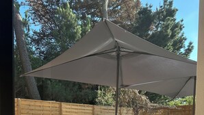 Un patio con mesa y sillas bajo un paraguas grande, cerca de una valla de madera, y un árbol al fondo.