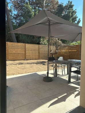A patio area with a table and chairs under a large umbrella, a wooden fence, and a tree in the background. - New house 155 m² /Seaside on foot (100 m)/4 beautiful bedrooms, garden, parking (Saint-Brevin-les-Pins)