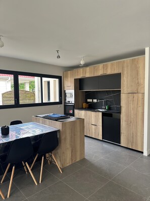 A modern kitchen with wooden cabinets, a black countertop, and a dining area with a table and chairs. - New house 155 m² /Seaside on foot (100 m)/4 beautiful bedrooms, garden, parking (Saint-Brevin-les-Pins)