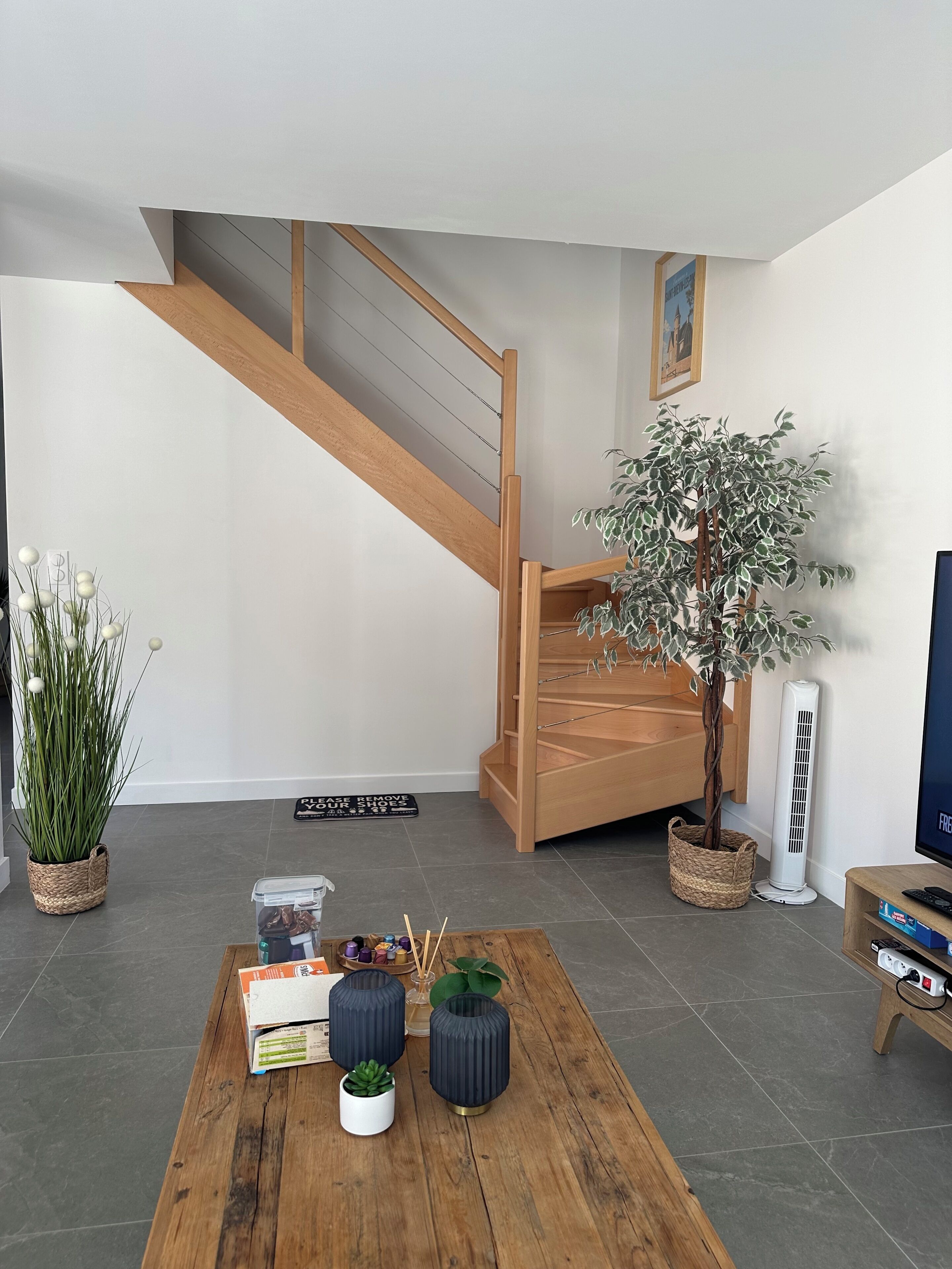 A modern living room with a wooden staircase, a large potted plant, a standing heater, and a wooden coffee table with books and decorative items.