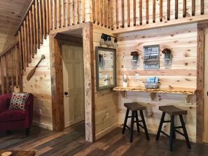 A wooden cabin interior with a staircase, a red sofa, two stools, and a mirror on the wall.