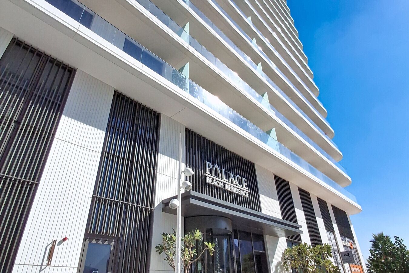 Et moderne hotel med et tydeligt skilt, der viser 'PALACE BEACH RESIDENCE', store vinduer og et rent arkitektonisk design.