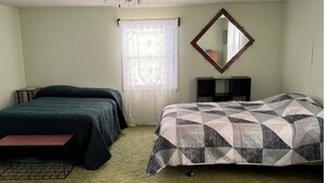 A bedroom with two beds, a window with a sheer curtain, a mirror on the wall, and a small bedside table.