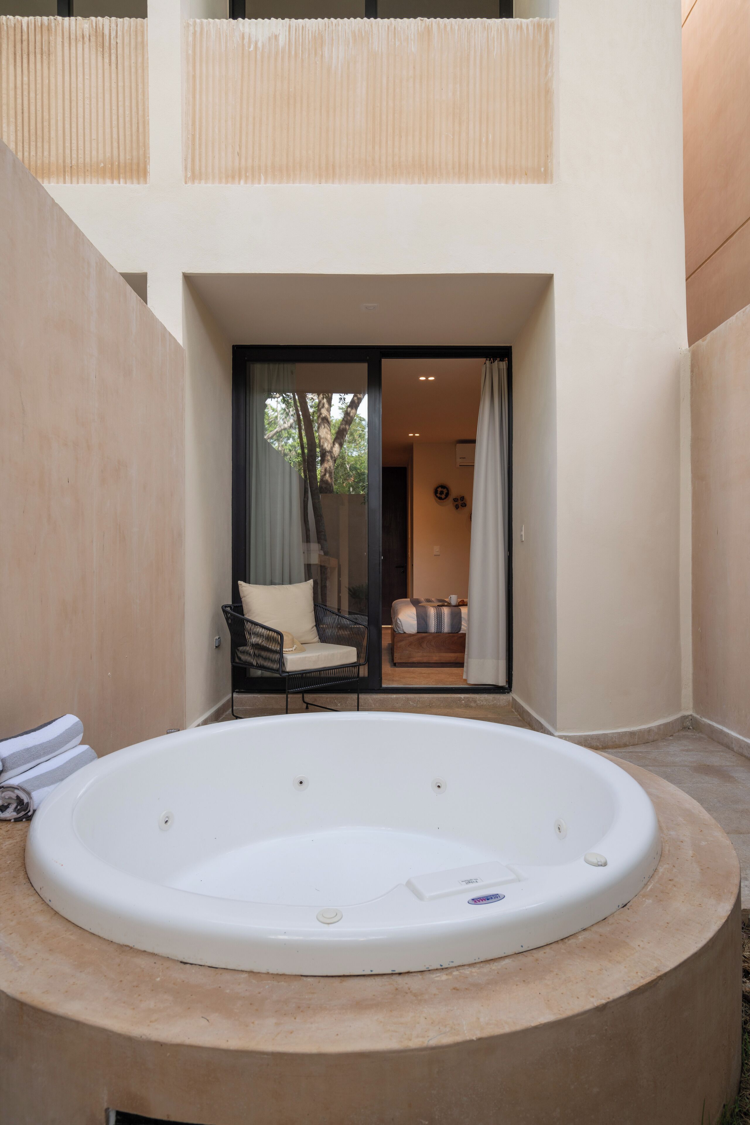 Un jacuzzi en un patio con una silla y un sofá, una puerta corrediza de vidrio que da a una habitación y una pared color beige.