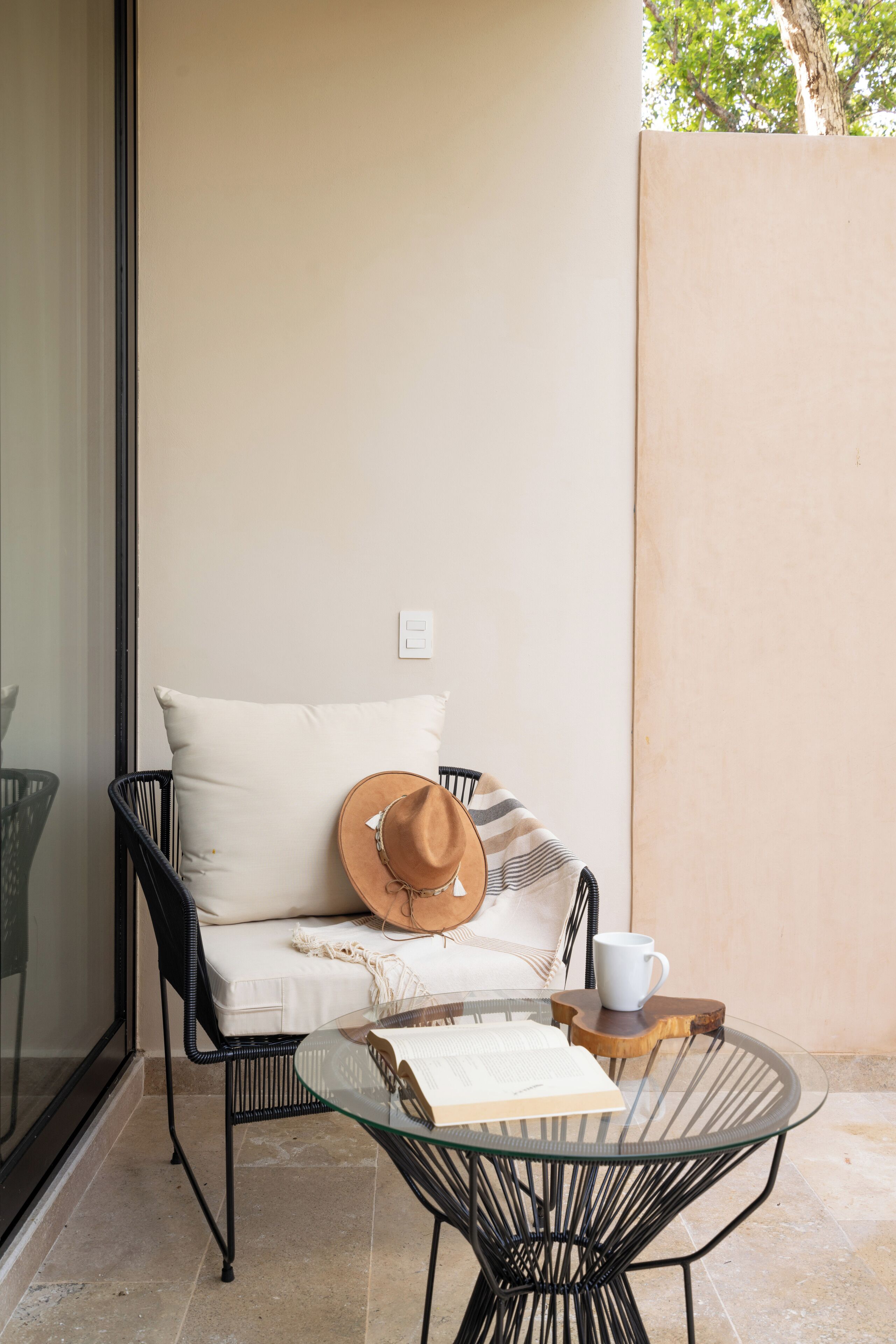 Un balcón con una mesa de vidrio, una silla de metal negro, un cojín blanco, un sombrero y una taza blanca.