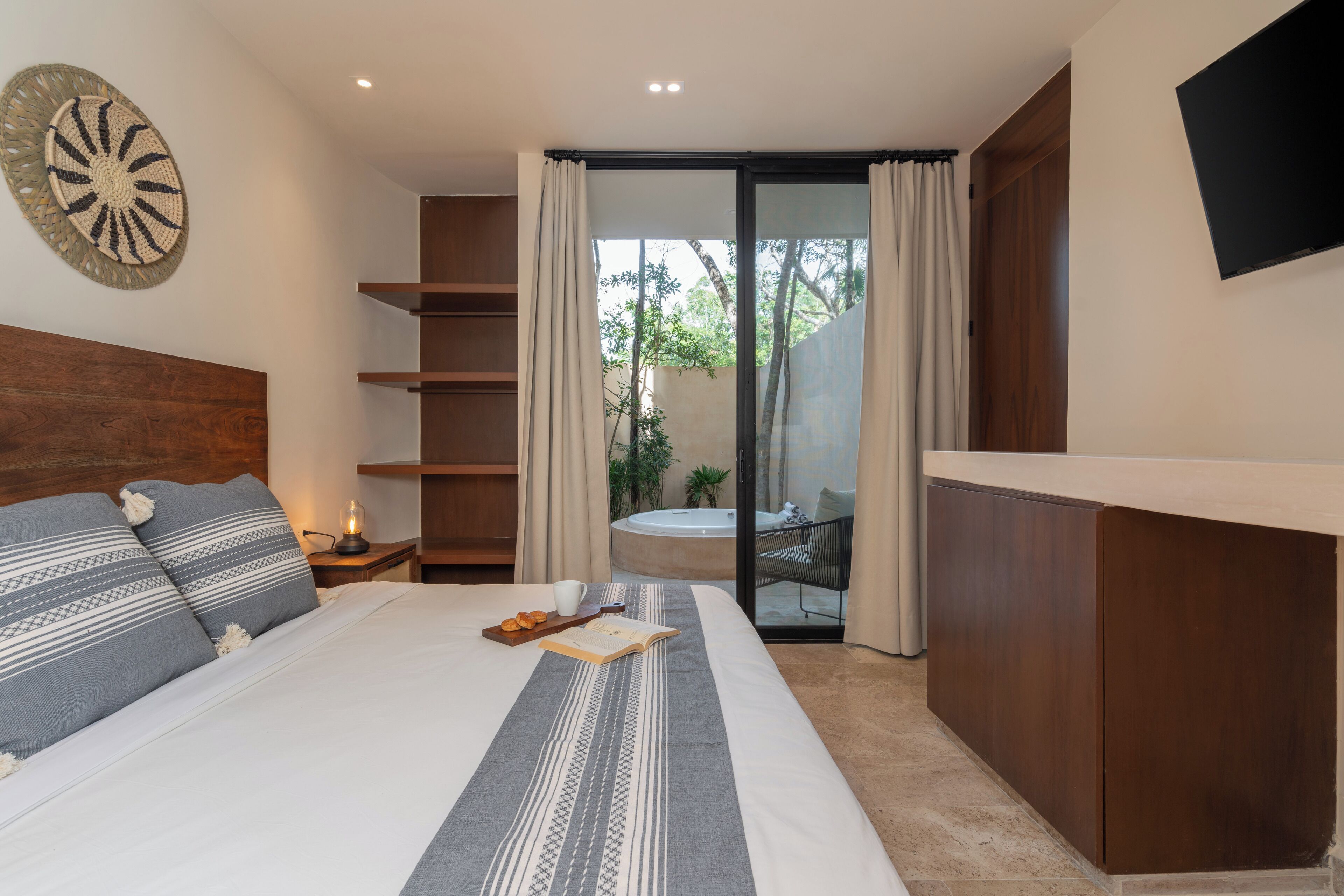 Una habitación de hotel moderna con una cama grande, cabecera de madera, televisor de pantalla plana, puerta corrediza de vidrio a un balcón y una pequeña mesa con un libro y una taza.