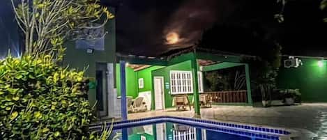 Une vue nocturne d’une maison avec une piscine, entourée de verdure et de carreaux de pierre.