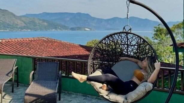 Iemand ontspant in een hangstoel op een balkon met uitzicht op zee en bergen.