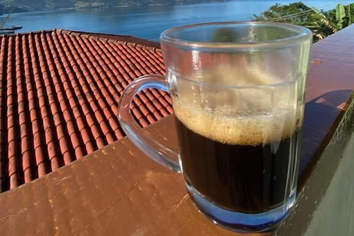 Une tasse en verre transparent contenant une boisson, posée sur une table devant un bâtiment au toit rouge, avec vue sur un lac et des montagnes lointaines.