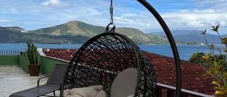 Una silla colgante con asiento y respaldo acolchados, una tumbona y una planta en maceta en un patio con vistas al agua y colinas.