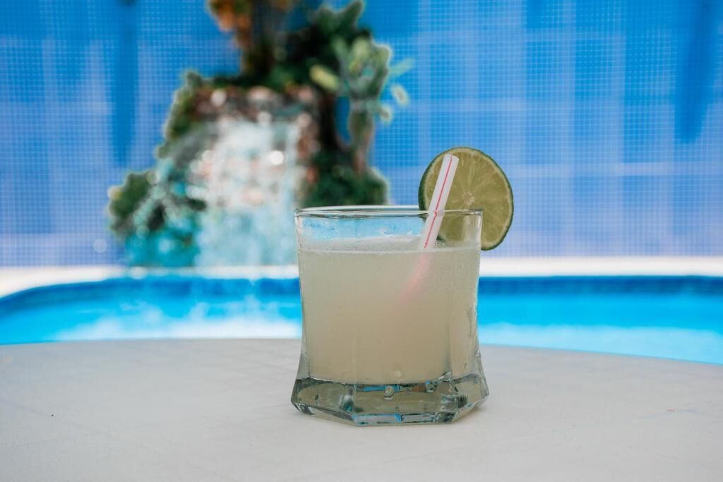 A glass of a light-colored beverage with a lime wedge and a straw, placed on a table near a pool.