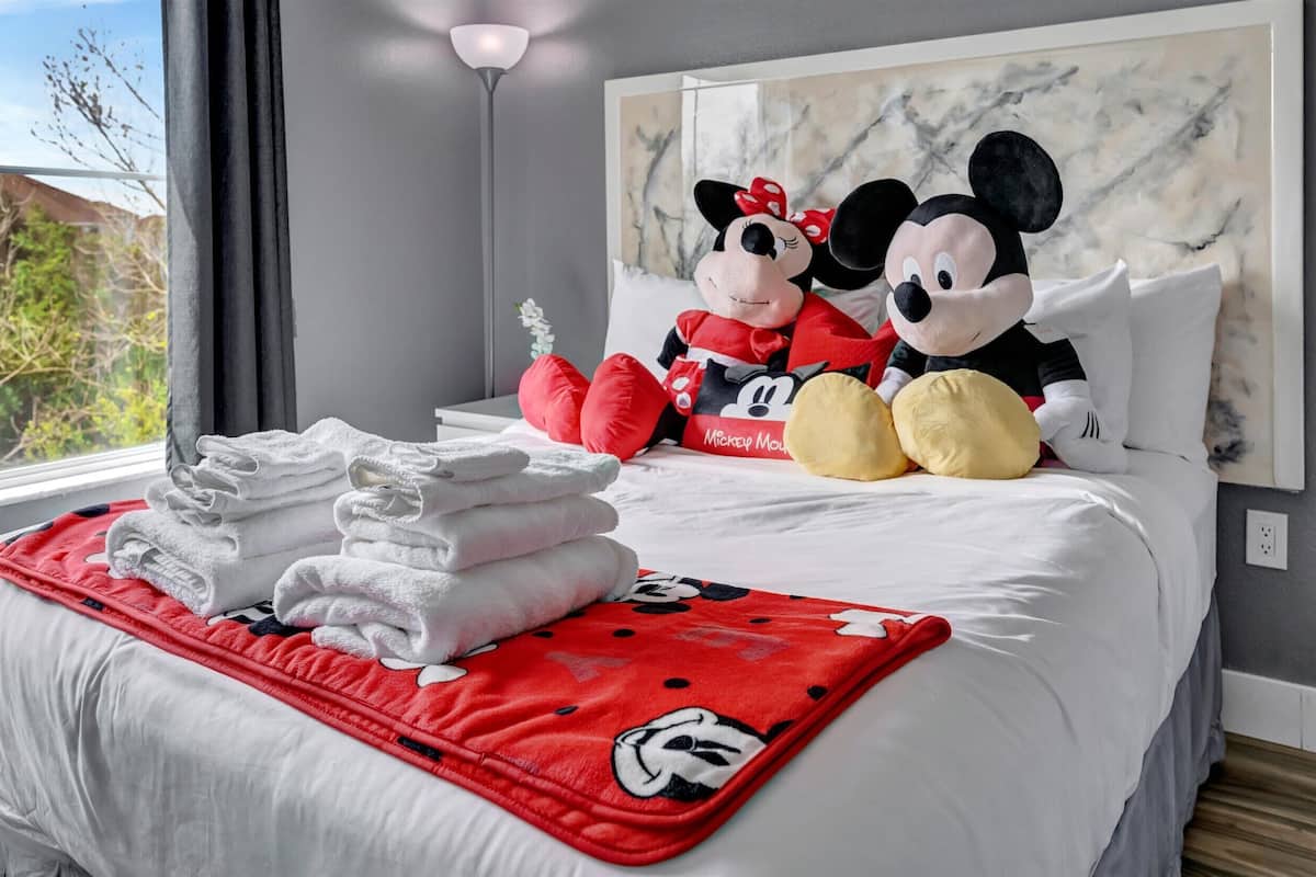 A neatly made bed with Mickey and Minnie Mouse plush toys, a red Mickey Mouse blanket, and folded towels on a white bedspread.