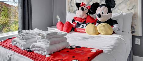 A neatly made bed with Mickey and Minnie Mouse plush toys, a red Mickey Mouse blanket, and folded towels on a white bedspread.