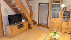 Een houten trap, een televisie op een houten kast, een glazen tafel met een afstandsbediening, en houten kasten met glazen deuren.