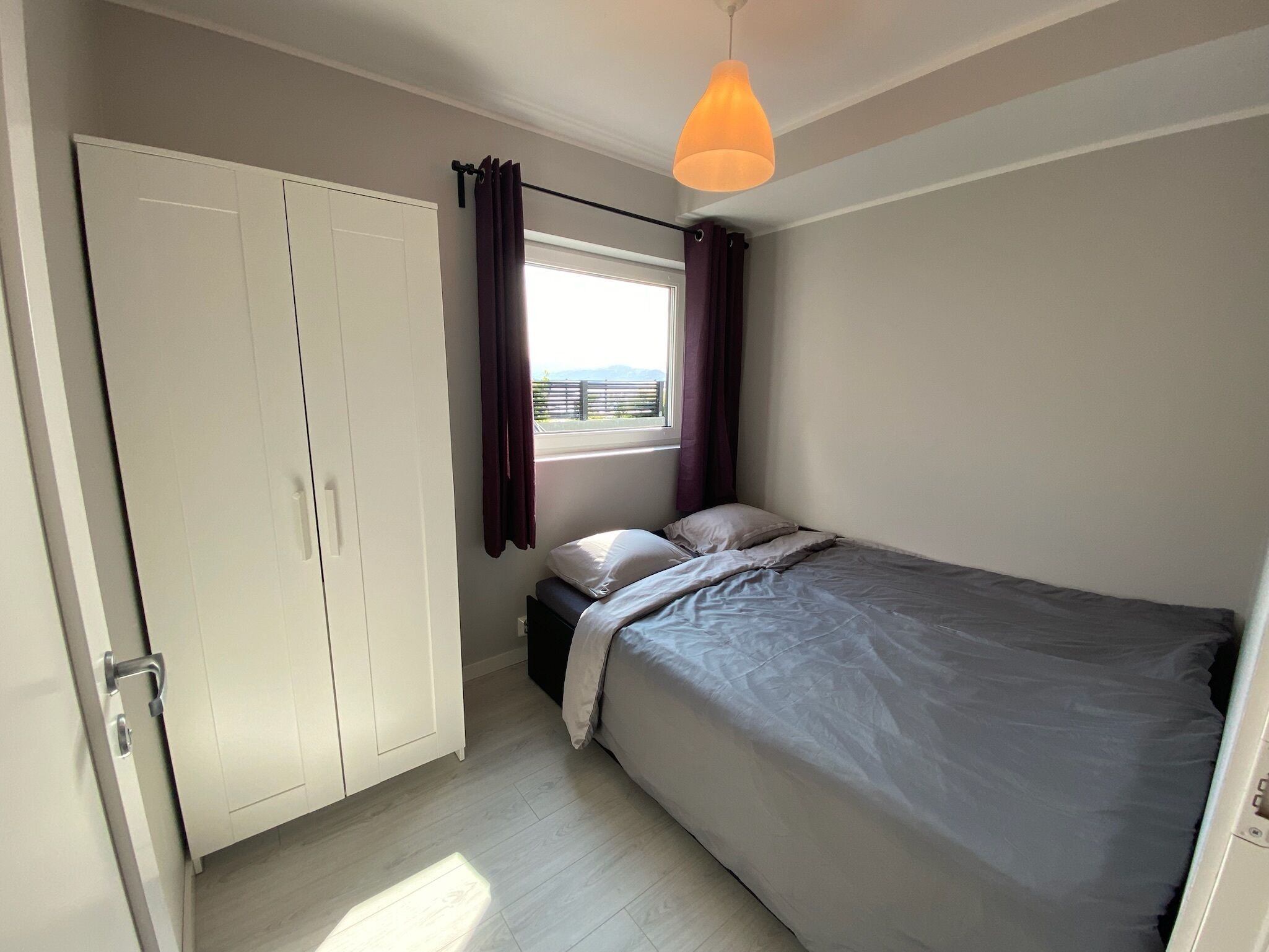 A bedroom with a bed, a wardrobe, a window with curtains, and a hanging light fixture.