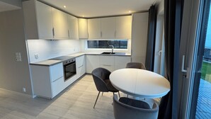 A modern kitchen with white cabinets, a built-in oven, and a window above the sink. There is a round table with two chairs and a view of the outdoors through a sliding door. - Beautiful ocean and fjord view short distance to Bergen (Vestland)