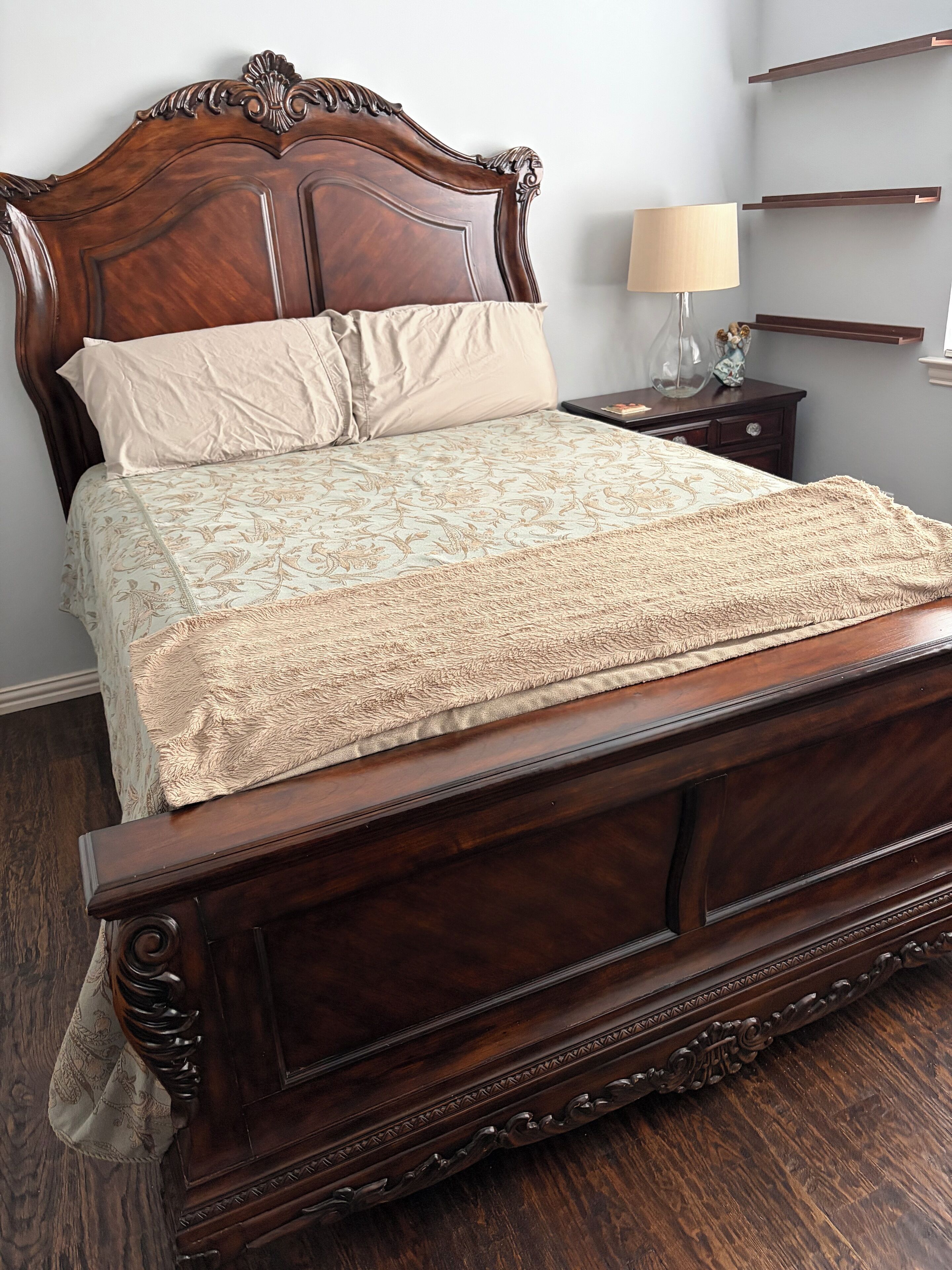 A wooden bed with a headboard, a bedside table with a lamp, and a wall-mounted shelf.