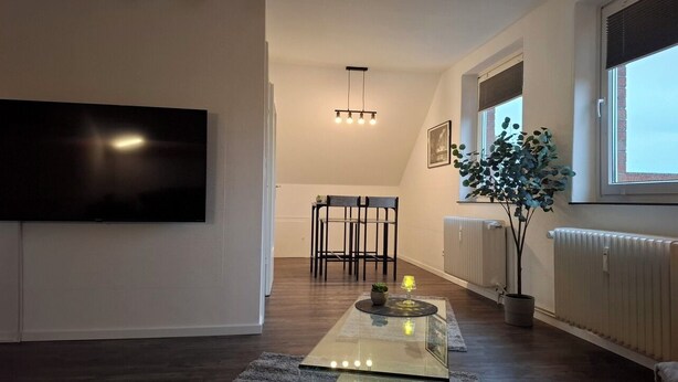 Uma sala de estar moderna com uma TV de tela plana, uma mesa de centro de vidro, uma poltrona, uma planta e um radiador.