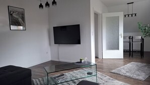 Uma sala de estar moderna com uma televisão de tela plana na parede, uma mesa de centro de vidro, um sofá preto e um tapete cinza.