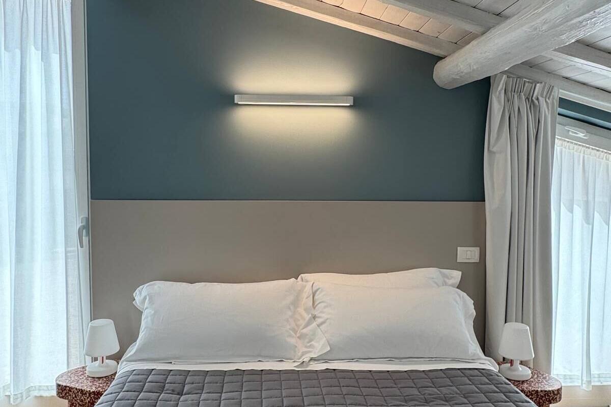 A bedroom with a bed, a bedside table, a wall-mounted light, and a window with curtains.