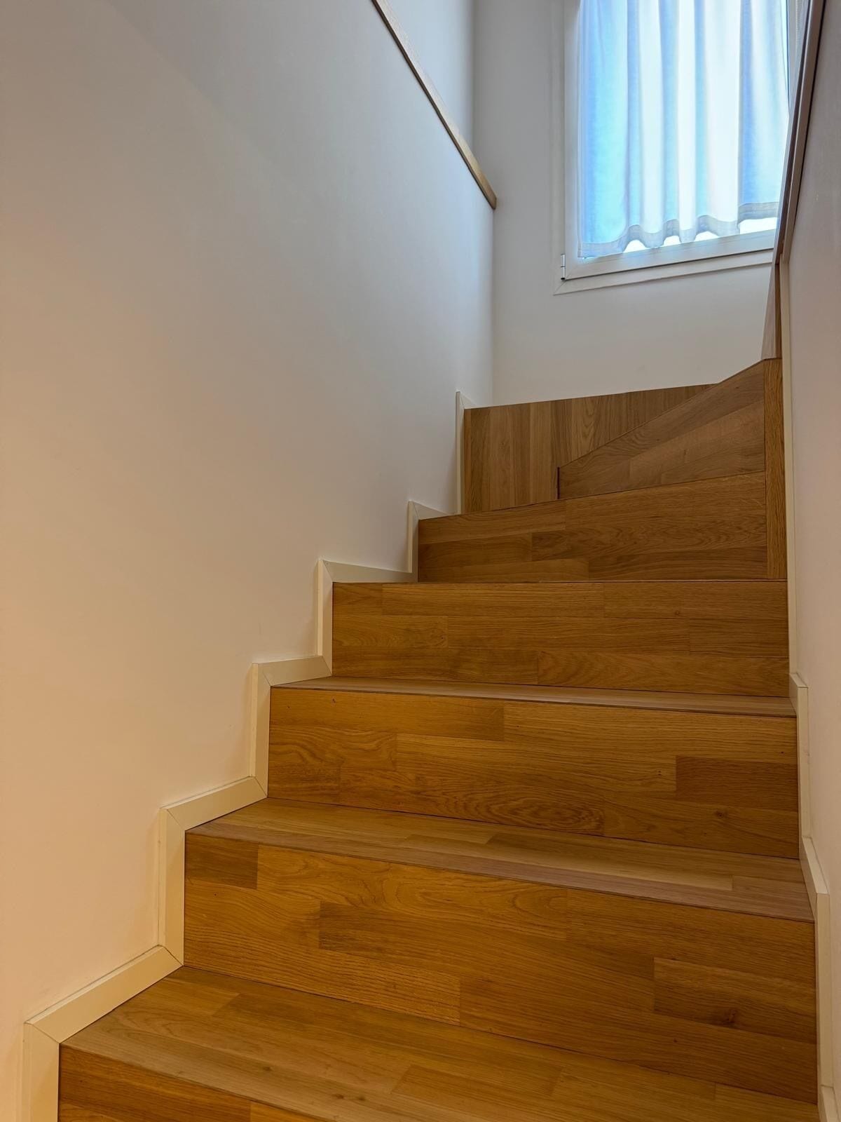 Un escalier en bois, des murs blancs et une fenêtre ornée de rideaux légers.