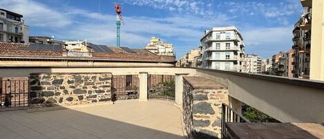 A rooftop terrace with tiled flooring, a stone wall, and a view of surrounding buildings.