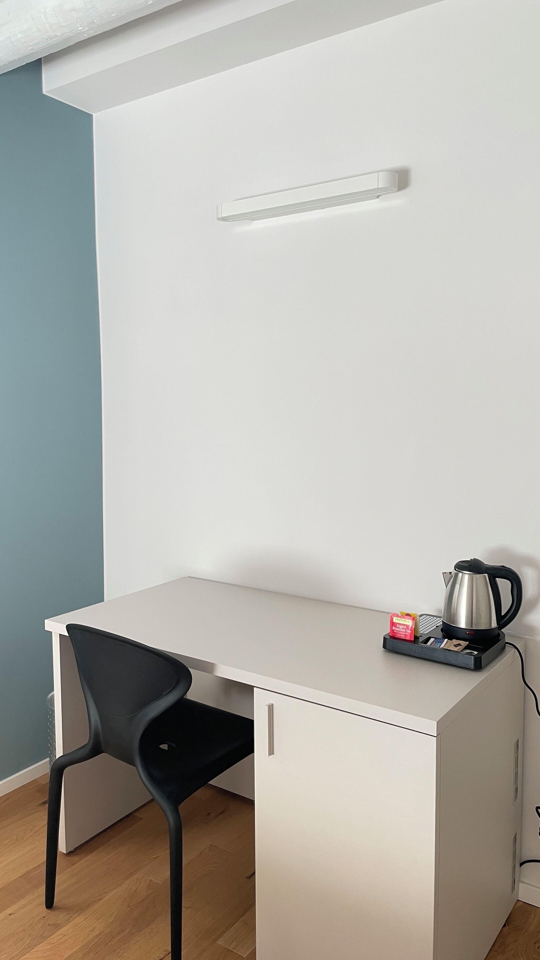 Un petit bureau blanc, une chaise noire, une bouilloire et un plateau avec une tasse et une boîte de mouchoirs.