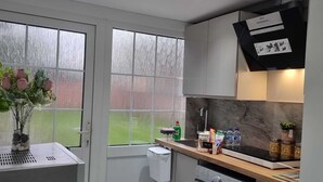 A compact kitchen with a washing machine, a sink, and a window with a view of a green area. - Brand New Studio Apartment (Barking)