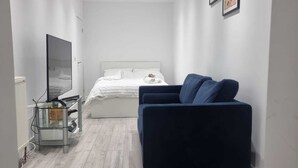 A narrow hotel room with a bed, a blue sofa, a TV, and a small table. - Brand New Studio Apartment (Barking)