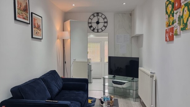 A modern living room with a dark blue sofa, a glass coffee table, a flat-screen TV on a glass stand, and a large wall clock. - Brand New Studio Apartment (Barking)