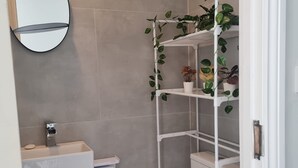 A modern bathroom with a toilet, sink, and mirror. There is a white shelf unit with plants and a toilet paper holder. - Brand New Studio Apartment (Barking)