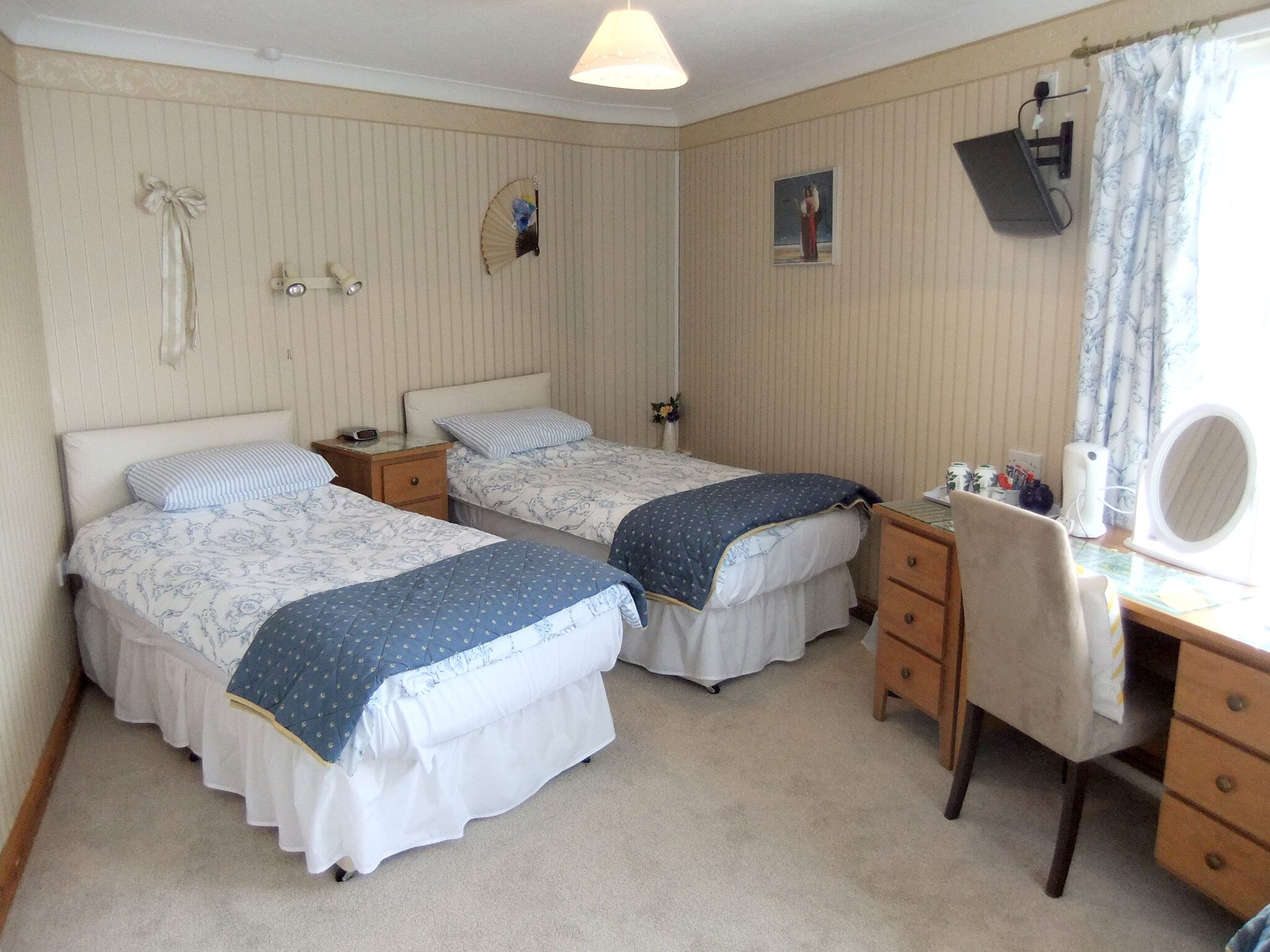 A bedroom with two single beds, a desk with a chair, a mirror, and a small bedside table with a lamp.