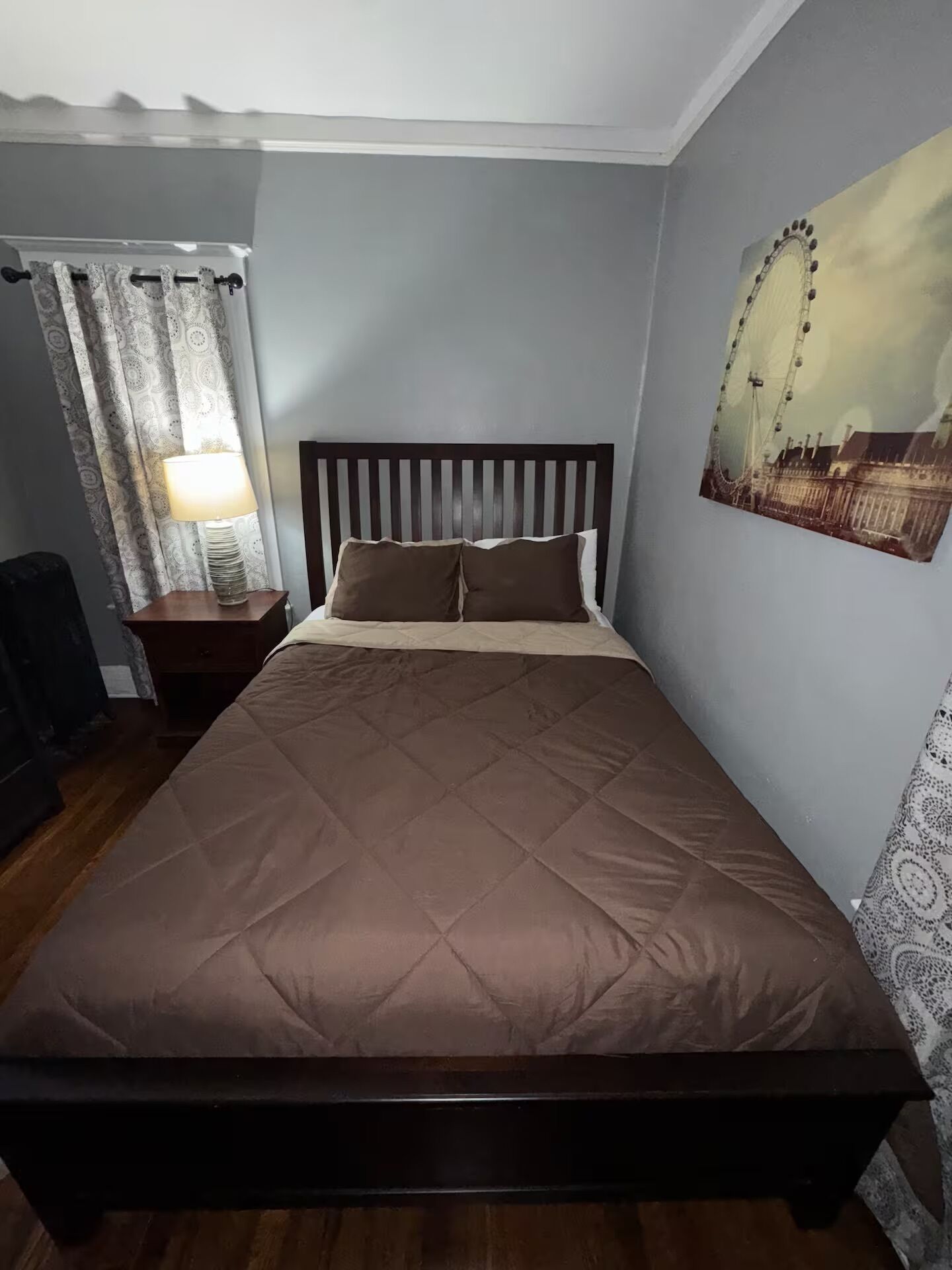 A bedroom with a large bed, a nightstand with a lamp, a wall-mounted picture of a Ferris wheel, and a patterned curtain.