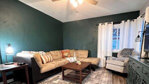 A living room with a dark teal wall, a beige sectional sofa, a wooden coffee table, a patterned rug, and a ceiling fan. - Historic Deereville Home Pet Friendly! (Dubuque)