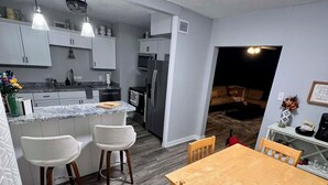 A modern kitchen with white cabinets, a granite countertop, and a dining area with a wooden table and chairs. - Historic Deereville Home Pet Friendly! (Dubuque)