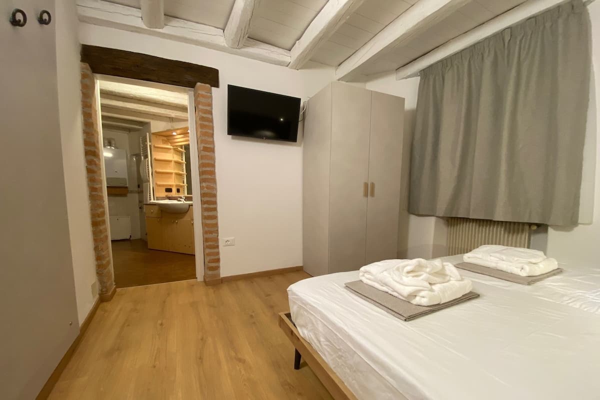 A bedroom with a wooden floor, a bed with white linens, a TV on the wall, and a bathroom visible through an open door.