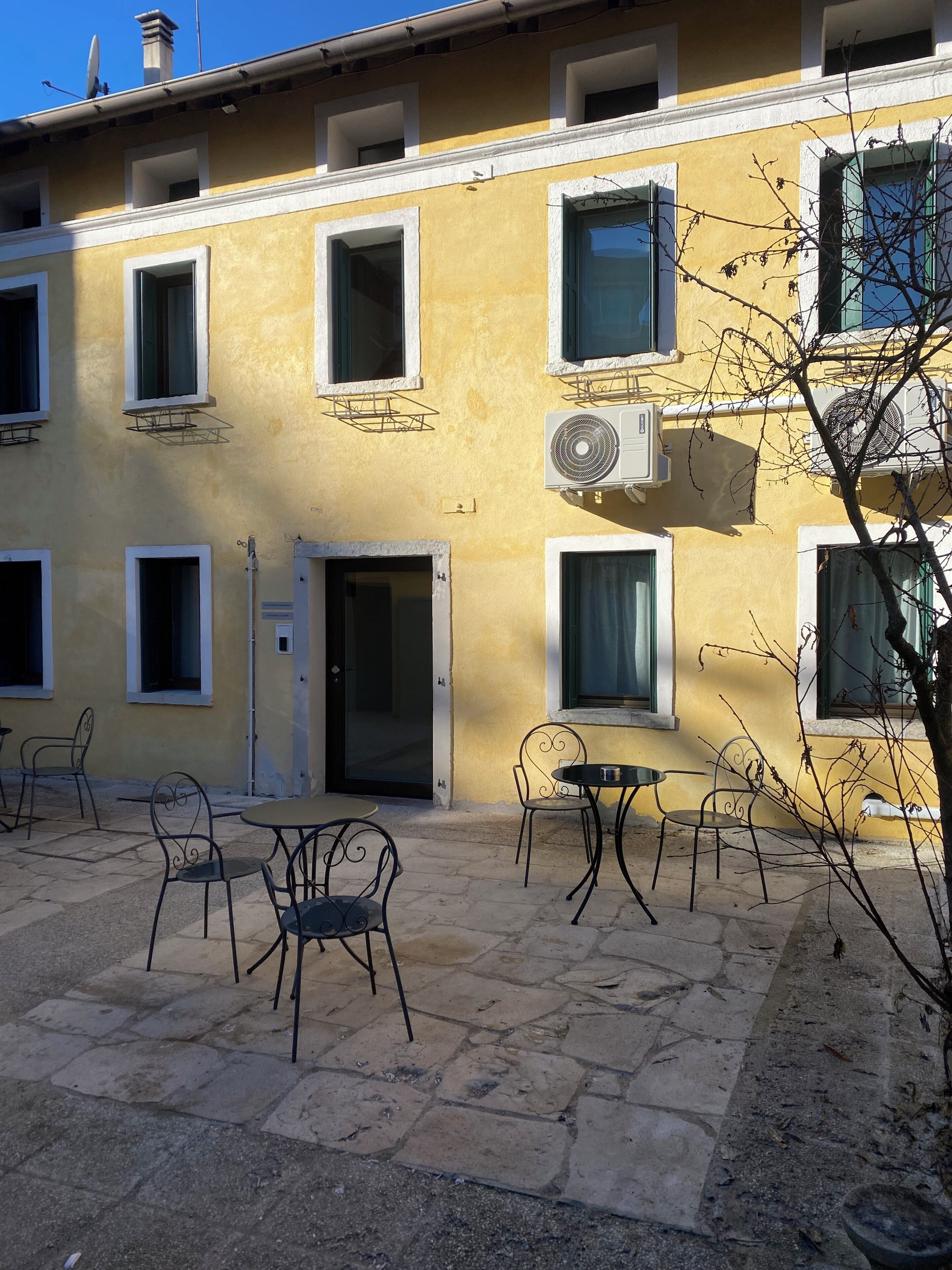 A building with multiple windows, an air conditioning unit, and outdoor seating with metal chairs and tables.