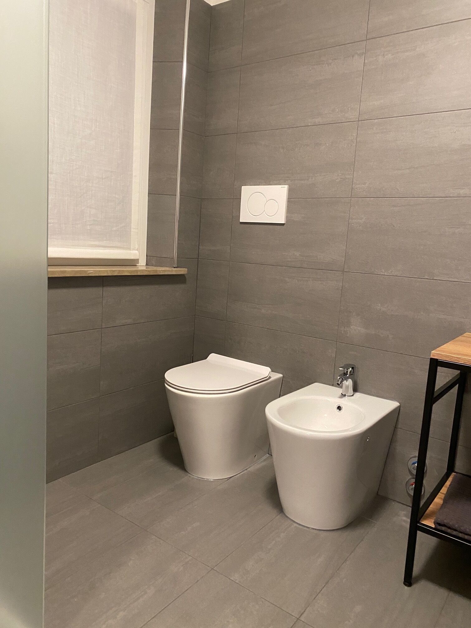 A small bathroom with a toilet and bidet, grey tiled walls, a window, and a wooden shelf.