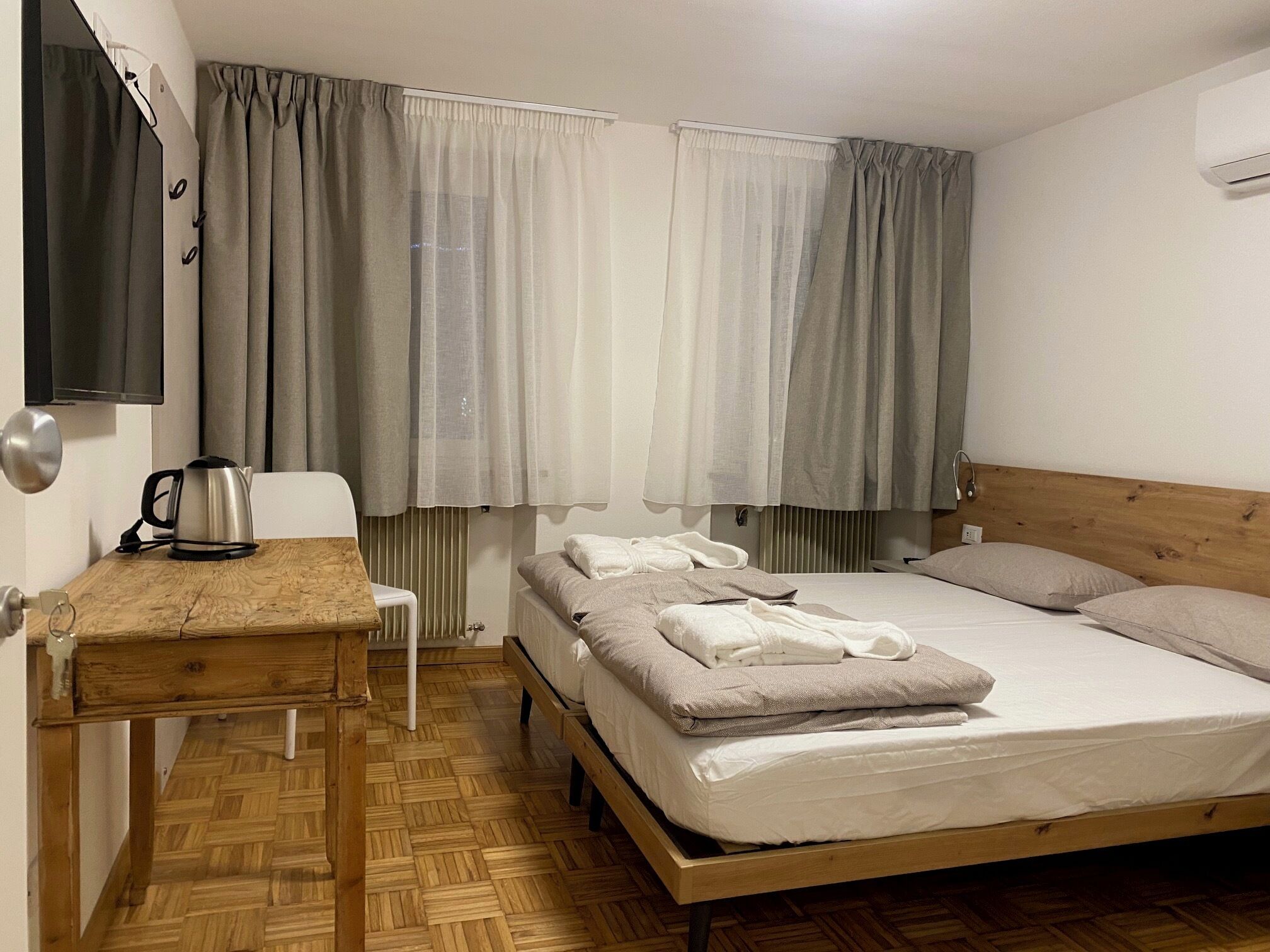 A bedroom with a wooden bed frame, a bedside table with a kettle, a chair, and a TV mounted on the wall.